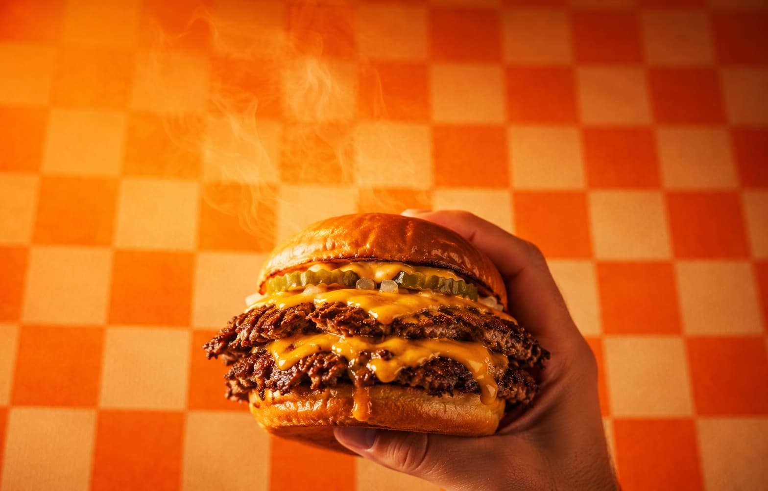 Smash burger Beldys fumant, tenu à la main devant une nappe damier orange